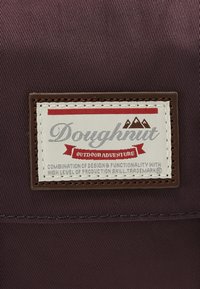 Brown fabric with a label featuring "Doughnut" in gray and red, mountains icon, and text about design functionality. Edge-stitched detail.