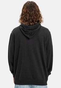 A person with curly hair faces away, wearing a black hoodie and light blue jeans, against a plain white background.