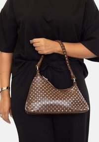 Bolso de cuero marrón con un acabado suave, adornado con lunares metálicos plateados y un asa trenzada. Diseño curvo y relajado.