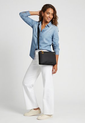 Kvinde smilende med den ene hånd bag hovedet, iført en denimskjorte, hvide bukser, beige sneakers og en sort crossbody-taske.