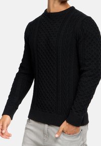 Zwarte gebreide trui met textuur in kabel- en wafelpatronen. Ronde halslijn en geribbelde manchetten. Gecombineerd met lichtgrijze broek.