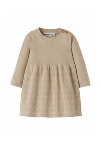 Beige neulemekko, jossa on teksturoitu yläosa, pitkät hihat ja liehuva hame, jossa on kukkakuvio helmassa. Kaksi puista nappia olkapäällä.