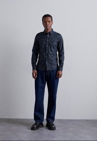 Camicia nera con bottoni, decorata con motivi colorati, abbinata a pantaloni di denim scuri e larghi, e scarpe nere, presentata su uno sfondo neutro.