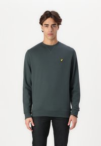 Felpa a maniche lunghe verde scuro con colletto tondo, polsini e orlo ribattuti. Presenta un piccolo logo di un uccellino giallo sul lato sinistro del petto.