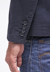 Mörkblå jackärm med fem svarta knappar längs mudden, med en strukturerad tyg och synliga syste detaljer.