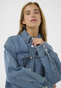 Veste en denim bleu clair avec des boutons-pression argentés, coupe ample, col pointu, poches avant et tissu texturé.