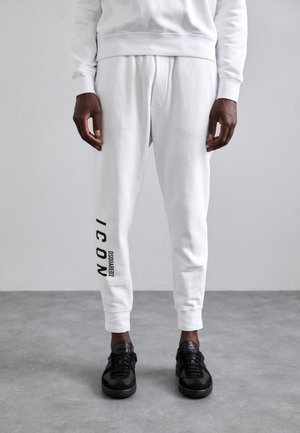 White sweatpants with a printed black "ICON" and brand logo on the side, elastic cuffs, paired with black sneakers. Smooth, casual fabric.