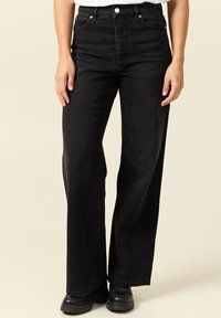 Persona vestida con jeans negros de pierna ancha de cintura alta y zapatos negros gruesos, de pie frente a un fondo claro y sencillo.