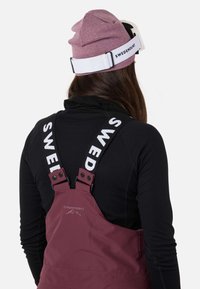 Maronde bib overalls med hvide logo-stropper, båret over en sort langærmet bluse. En grå beanie sidder på hovedet, med skibriller.