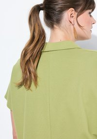 Blusa verde con una finitura testurizzata, maniche corte e colletto. Presenta una cucitura sul retro e un modello con coda di cavallo e orecchini.
