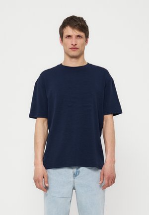 Homme portant une chemise bleu marine texturée à manches courtes et un jean bleu clair, debout devant un fond blanc uni.
