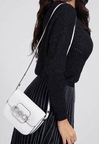 Bílá kožená crossbody taška se strukturovaným tvarem, stříbrným kováním a řetízkovým akcentem. Spárována s černým texturovaným svetr a plisovanou sukni.