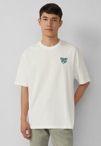 Jeune homme portant un t-shirt blanc avec le texte "Sortie Scolaire" sur le côté gauche de la poitrine, se tenant devant un fond gris clair uni.