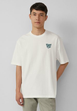 Jonge man in een wit T-shirt met de tekst "Field Trip" op de linkerkant van de borst, staand tegen een effen lichtgrijze achtergrond.