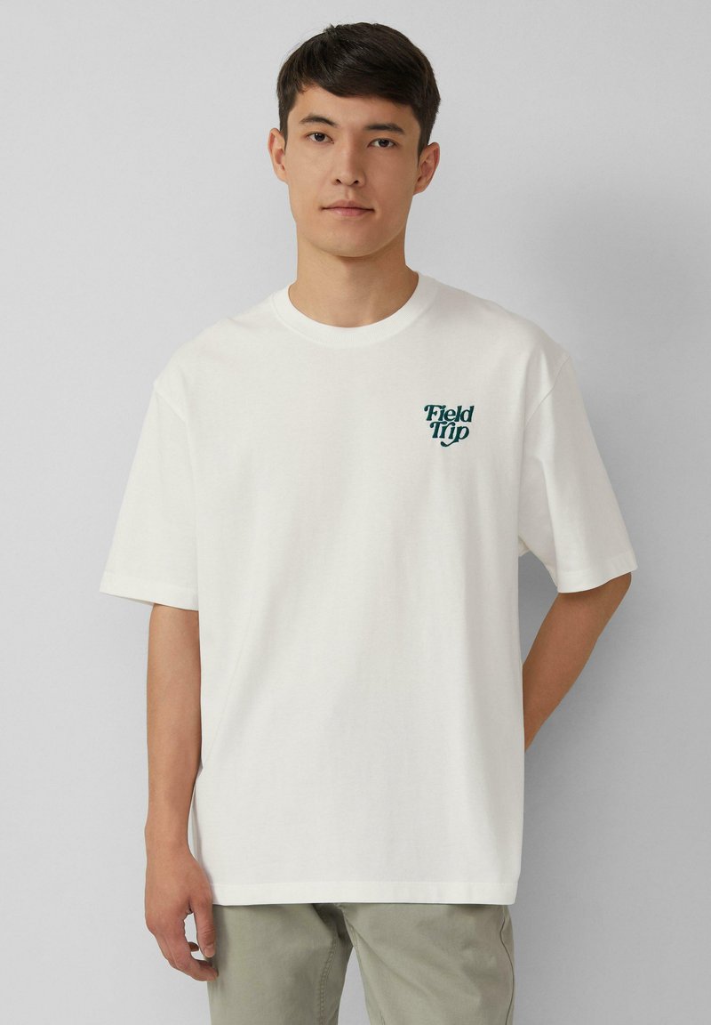 Jeune homme portant un t-shirt blanc avec le texte "Sortie Scolaire" sur le côté gauche de la poitrine, se tenant devant un fond gris clair uni.