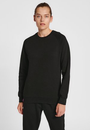 Jeune femme aux cheveux attachés, vêtue d'un sweat-shirt noir uni à manches longues et d'un pantalon noir, debout devant un fond blanc.