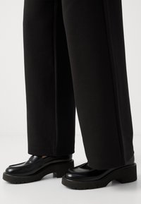 Černé kožené platformové mokasíny s hladkým povrchem a tvarovaným designem, doplněné širokými černými látkovými kalhotami. Masivní podrážky.