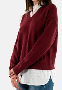 Maglione borgogna con scollatura a V, polsini e fondo a coste; indossato sopra una camicia bianca con bottoni caratterizzata da sottili pois.