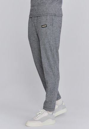 Gray herringbone sweatpants, fitted design with side pockets, black label, paired with white mesh sneakers, ribbed cuffs visible.