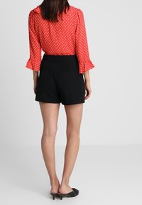 Blusa rossa a pois con maniche a volant, abbinata a pantaloni corti neri a vita alta. L'outfit è completato da scarpe nere con il tacco.