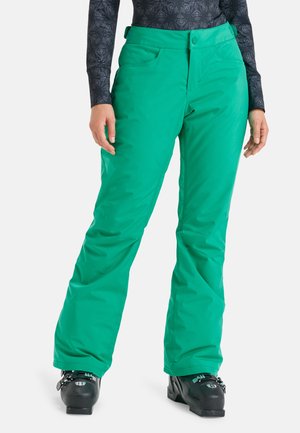 Zielone spodnie narciarskie noszone z czarnymi butami narciarskimi, połączone z ciemnym wzorzystym topem z długim rękawem, pokazane od talii do stóp.