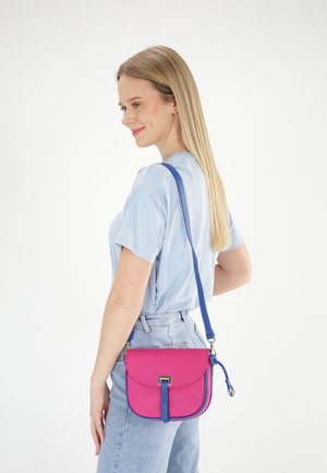 Lyserød læder crossbody taske med en blå rem, der har en rund form, knaplukning og tekstureret overflade. Båret på siden.