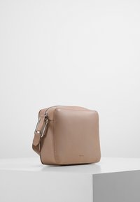 Piccola borsa a tracolla in pelle beige con fibbia e cerniera argentate, design minimalista, posizionata su una superficie bianca contro uno sfondo grigio chiaro.