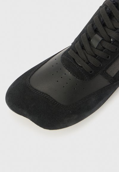 Crne atletske tenisice s kombinacijom teksturirane antilop kože i glatke kože. Uključuju perforacije i vezanje na vrhu.