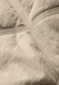 Beige, geblokt stof met zichtbare stiksels in een X-patroon, met een zachte textuur en een glad oppervlak. Close-upweergave benadrukt de details.