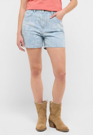 Frau trägt hellblaue Jeansshorts, korallenfarbenes Oberteil und hellbraune Wildleder-Stiefeletten mit Blockabsätzen, steht mit einer Hand in der Tasche.