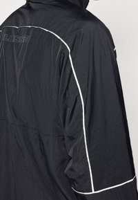 Osoba nosząca czarną lekką kurtkę z białym przeszyciem wzdłuż szwów i subtelnym napisem z nazwą marki na górnej części pleców.