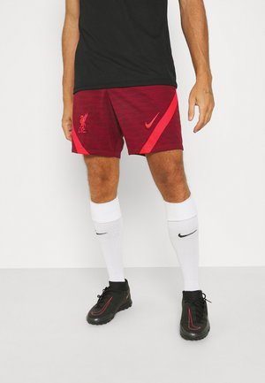 Un mannequin masculin porte un t-shirt noir, un short bordeaux de Liverpool FC avec logo, des chaussettes blanches et des chaussures Nike noires. Fond clair uniforme.