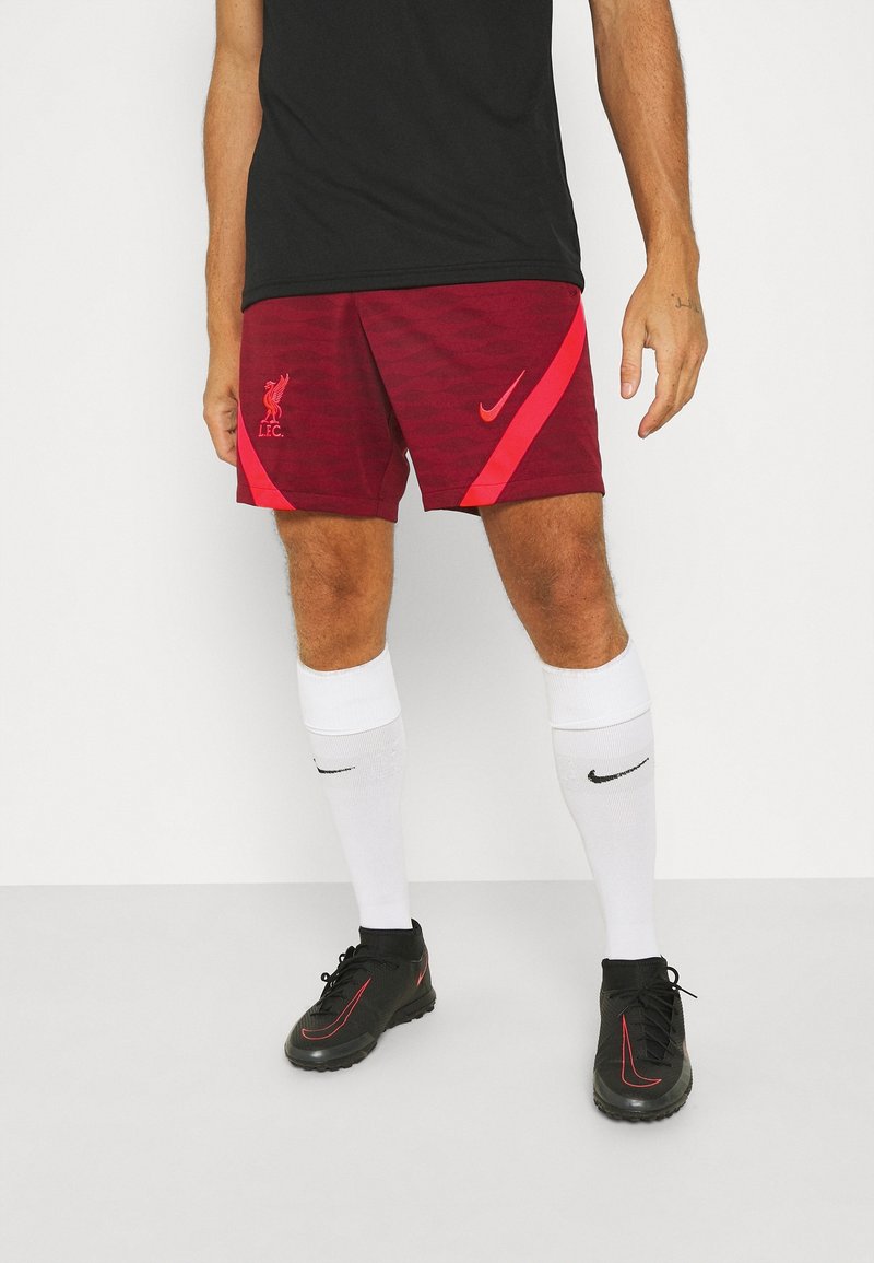 Miesmalle mulla on päällä musta t-paita, Liverpool FC:n maroonit shortsit logolla, valkoiset sukat ja mustat Nike-kengät. Yksinkertainen vaalea tausta.