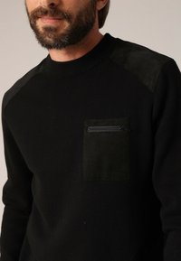 Zwarte gebreide trui met een gestructureerd zwart patchvak met rits en suède-achtige schouderaccenten. Ronde halsontwerp met geribbelde manchetten.