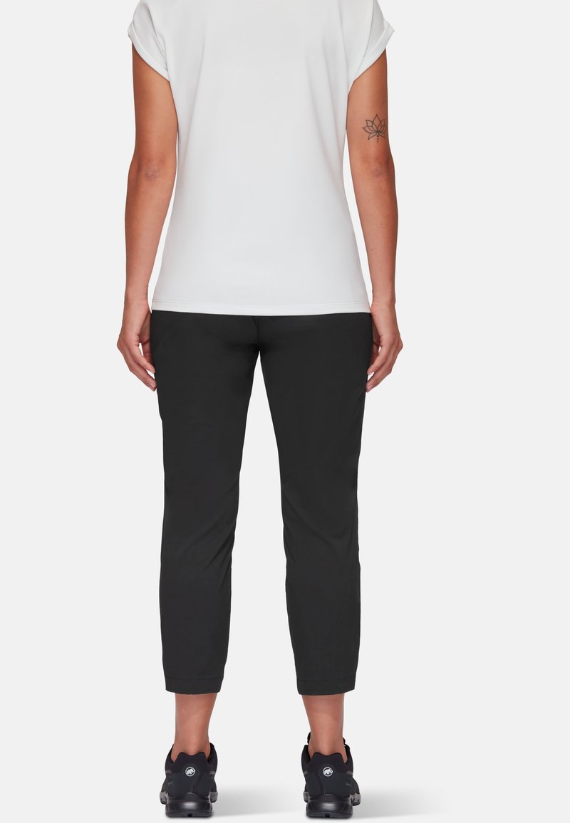 Czarne dopasowane spodnie o gładkiej fakturze, w połączeniu z białą bluzką z krótkim rękawem; widoczny tatuaż na nodze i czarne buty sportowe.