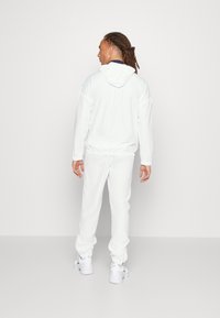 Baltas treniruočių kostiumas, turintis gobtuvą ir elastines rankoves. Sklandus audinys, laisvo kirpimo. Suderintas su baltais sportbačiais.