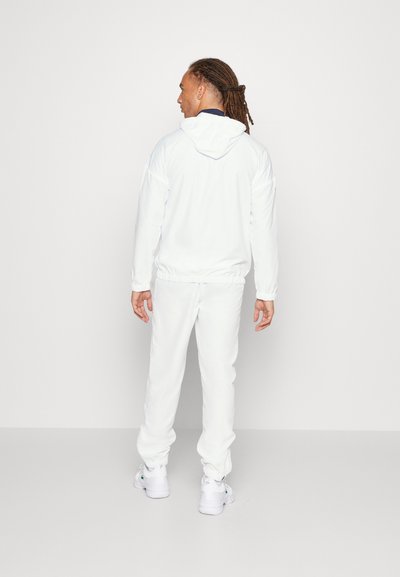 Бял tracksuit с качулка и еластични маншети. Гладка материя с свободно прилепване. Съчетава се с бели маратонки.