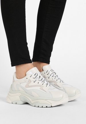 White chunky sneakers with black and white laces worn with black fitted pants on a plain white background.