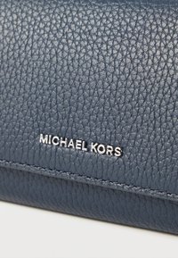 Tamnoplavi kožni novčanik s teksturiranim završetkom, sa srebrnim natpisom "MICHAEL KORS" na dnu i ušivenim detaljima duž rubova.
