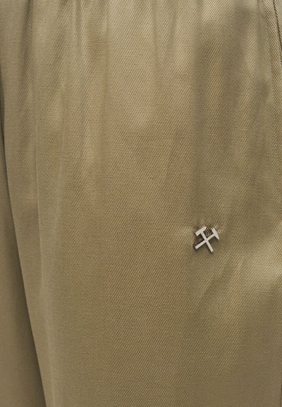 Κοντινό πλάνο μπεζ υφάσματος με μια μικρή ασημένια καρφίτσα σε σχήμα σταυρωτών σφυριών, τοποθετημένη κοντά στην κάτω δεξιά γωνία.