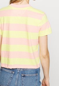 Persona vestida con una camiseta de rayas horizontales en tonos rosa pastel y amarillo, y unos pantalones de mezclilla azul de cintura alta, vista desde atrás.