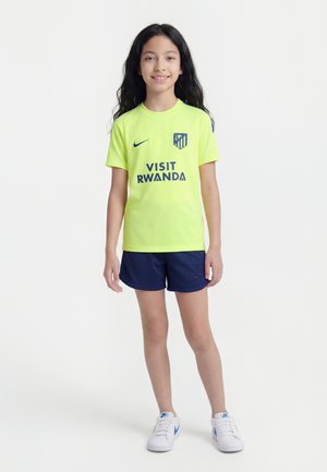 Jeune fille debout, portant un maillot de football jaune fluo d’Atletico Madrid, un short bleu marine et des baskets blanches, sur un fond blanc uni.