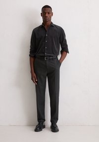 Un homme se tient contre un mur blanc, portant une chemise noire à manches retroussées, un pantalon noir ajusté, une ceinture noire et des chaussures de ville noires.