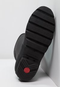 Botas de goma negras con una suela texturizada, que presenta surcos profundos para mayor tracción y un acento circular rojo en el talón. Nombre de la marca "HUNTER" visible.