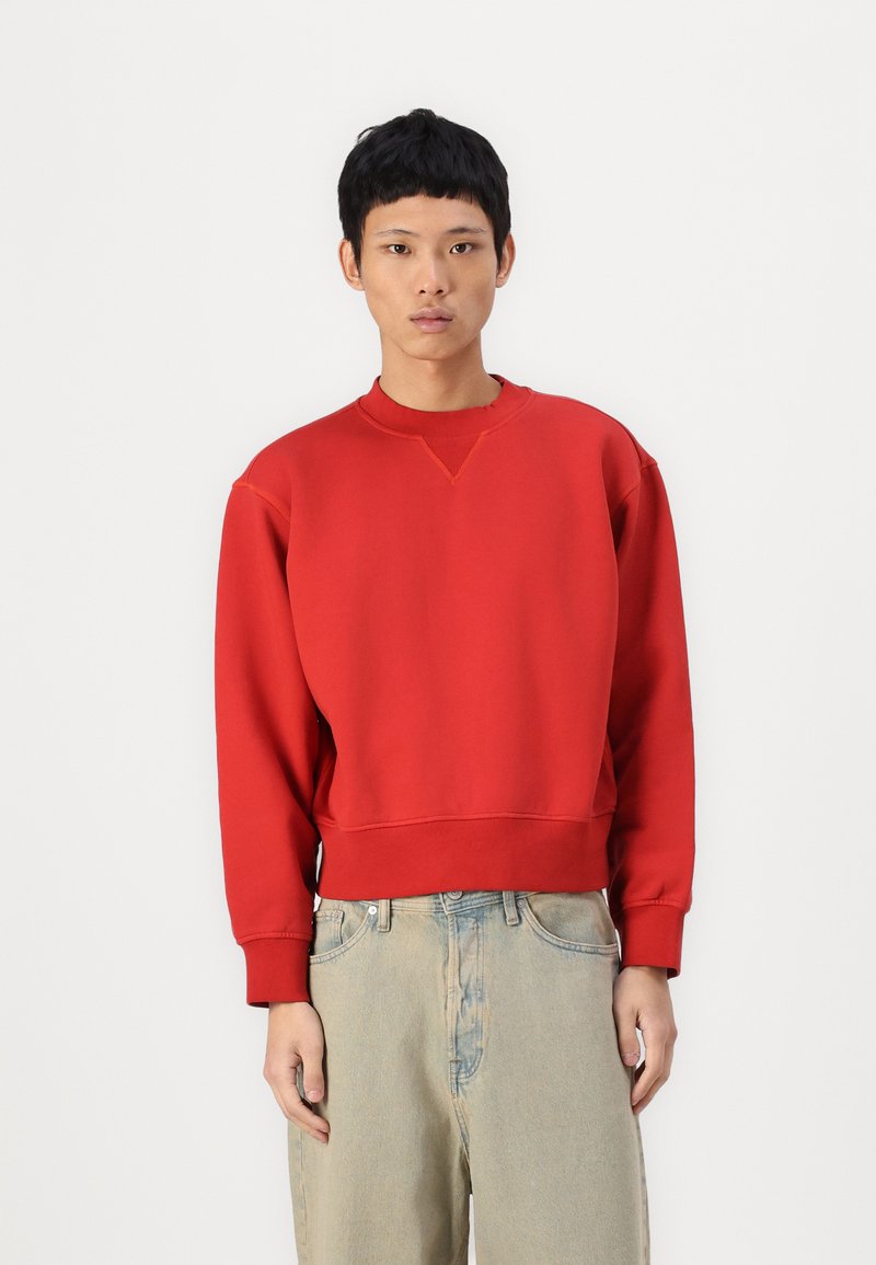 Rød hettegenser med rund hals, oversized passform, ribbede mansjetter og strukturert stoff. Kombinert med lyseblå, litt slitte jeans.