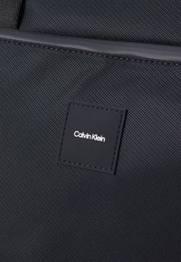 Juodas audinė maišelis su tekstūruota paviršiumi ir stačiakampiu Calvin Klein logotipo ženkleliu, kuris išryškintas. Ypatybės: tvirtas siuvinėjimas.