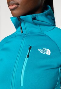 Giacca leggera teal con zip e colletto alto, dotata di tasca sul petto con zip e un logo bianco prominente sul davanti.