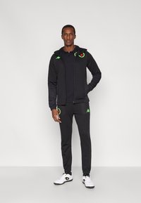 Čierna mikina na zips s kapucňou a zodpovedajúce tepláky. Má zelené logo a vrecká. Nosené s bielymi teniskami.