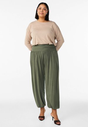 Žena stojaca s rukami za chrbtom, oblečená v béžovom tričku s dlhým rukávom, voľných olivovo zelených nohaviciach a čiernych sandáloch na opätku na bielom pozadí.