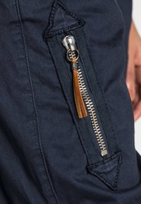 ciemnoniebieska tkanina z wyraźnym srebrnym zamkiem błyskawicznym z brązowym skórzanym uchwytem, ozdobiona kontrastowym przeszyciem i trójkątną łatą nad zamkiem.